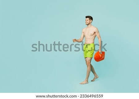 Full body side view young lifeguard man wear green shorts swimsuit relax near hotel pool hold lifebuoy walk go look aside isolated on plain blue background. Summer vacation sea rest sun tan concept