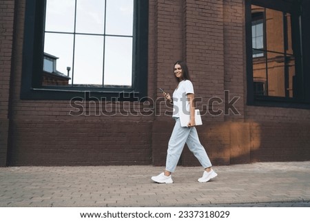Full body side view of young female student in white shirt walking on pavement near modern building and browsing smartphone while looking at camera