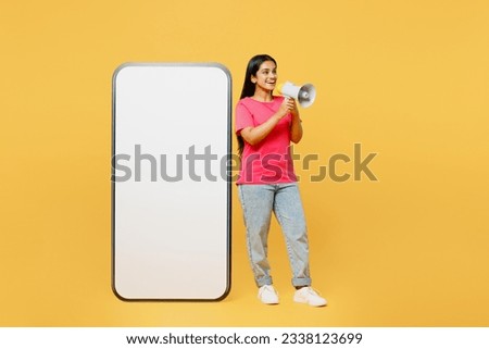 Full body side view young fun Indian woman wear pink t-shirt casual clothes big huge blank screen mobile cell phone smartphone with area scream in megaphone isolated on plain yellow background studio