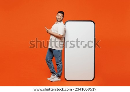Full body side view young Indian man wears white t-shirt casual clothes big huge blank screen mobile cell phone with area using smartphone isolated on plain orange red background. Lifestyle concept