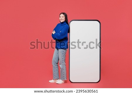 Full body side view young woman of Asian ethnicity wears blue sweater casual clothes stand near big huge blank screen mobile cell phone smartphone with area isolated on plain pastel pink background