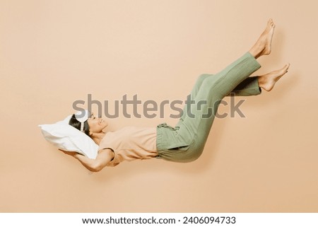 Full body side view young calm Latin woman wears pyjamas jam sleep eye mask rest relax at home fly up hover over air fall down on pillow isolated on plain beige background. Good mood night nap concept