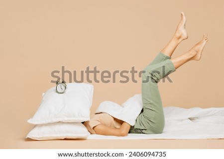 Full body side view young woman wears pyjamas jam sleep eye mask rest relax at home under duvet cover head with pillow alarm clock on it isolated on plain beige background. Bad mood night nap concept