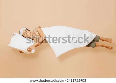 Full body side view young Latin woman wears pyjamas jam sleep eye mask rest relax at home fly fall hover over air on pillow under duvet isolated on plain beige background. Good mood night nap concept