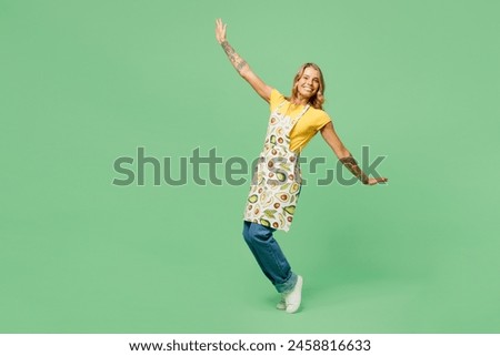 Full body side view young housewife housekeeper chef cook baker woman wear apron yellow t-shirt stand on toes lean back with outstretched hands isolated on plain green background. Cooking food concept