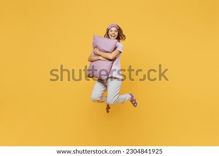 Full body sideways fun young woman she wears purple pyjamas jam sleep eye mask rest relax at home jump high hold in hand pillow isolated on plain yellow background studio portrait. Night nap concept