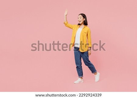 Full body sideways happy young woman of Asian ethnicity wear yellow shirt white t-shirt looking camera walking going stroll waving hand isolated on plain pastel light pink background studio portrait