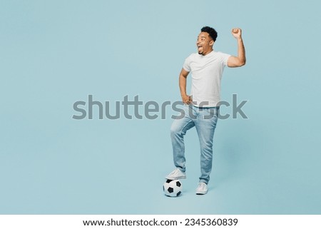 Full body sideways happy young man fan wear t-shirt cheer up support football sport team put leg on soccer ball watch tv live stream do winner gesture isolated on plain pastel blue color background