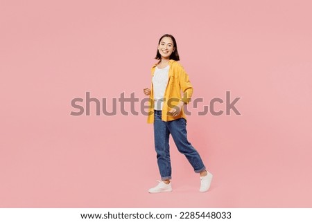 Full body sideways smiling happy young woman of Asian ethnicity wear yellow shirt white t-shirt looking camera walking going strolling isolated on plain pastel light pink background studio portrait