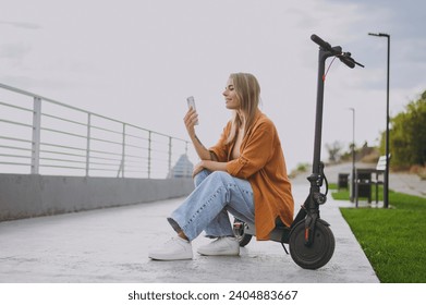 Full body sideways young fun cool woman wear orange shirt casual clothes sit on e-scooter talk on mobile cell phone walk resting relax in spring city park outdoors on nature. Urban lifestyle concept - Powered by Shutterstock