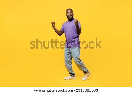 Full body sideways young man of African American ethnicity wears casual clothes purple t-shirt walk go do winner gesture celebrate clench fists say yes look camera isolated on plain yellow background