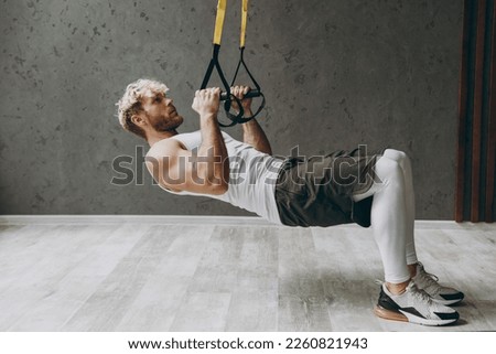 Full body sideways young strong sporty athletic sportsman man wearing white tank shirt black shorts doing exercises with trx loops looking camera warm up training indoor at gym. Workout sport concept