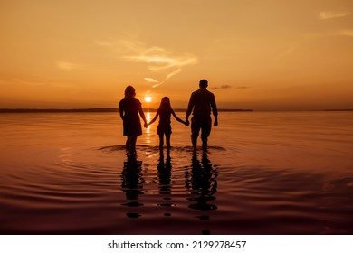 Full Body Silhouette Of An Unrecognizable Family Holding Hands And Walking With Their Backs To The Camera On The Water Of A River, During Sunset, Copy Space, Toned Image