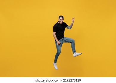 Full Body Singer Young Bearded Tattooed Man 20s He Wears Casual Black T-shirt Cap Jump High Play Guitar Hands Gesture Isolated On Plain Yellow Wall Background Studio Portrait. Tattoo Translate Fun.