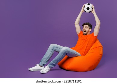 Full body size excited young fan man wear orange t-shirt cheer up support football sport team hold soccer ball above head watch tv live stream sit in bag chair isolated on plain dark purple background - Powered by Shutterstock