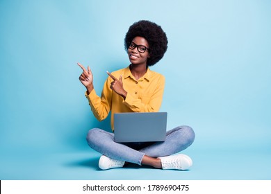 Full body size photo of black skin big volume hairstyle woman sit floor crossed legs hold netbook direct fingers empty space wear specs jeans yellow shirt isolated blue color background - Powered by Shutterstock