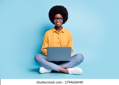 Full Body Size Photo Of Black Skin Big Volume Hairstyle Woman Sit Floor Hold Netbook Shiny Smiling Cozy Workplace Coding Website Wear Specs Jeans Yellow Shirt Isolated Blue Color Background