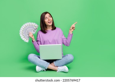 Full Body Size Photo Of Young Woman Youtube Blogger Rich Sitting With Laptop Hold Cash Dollars Interested Finger Pointing Empty Space Isolated On Green Color Background