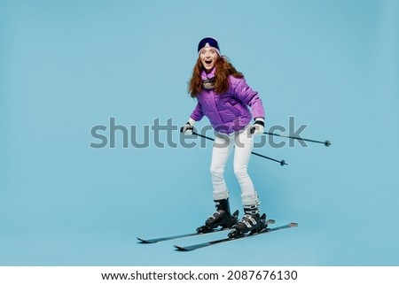 Full body skier amazed happy fun cool woman 20s wearing warm purple padded windbreaker jacket ski goggles mask spend extreme weekend in mountains look camera isolated on plain blue background studio.