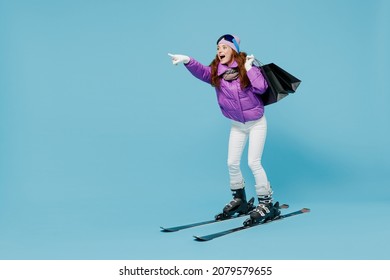 Full Body Skier Woman In Warm Purple Jacket Ski Goggles Mask Spend Weekend In Mountains Holding Package Bags With Purchases After Shopping Point Finger Aside Isolated On Plain Blue Background Studio.