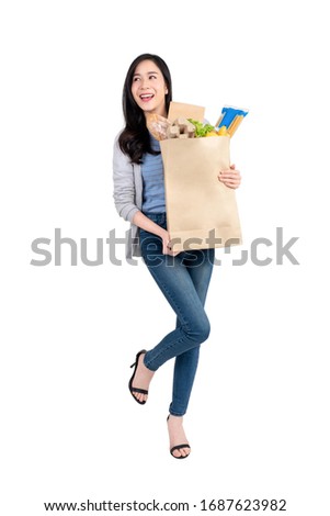 Full body of smiling Asian woman holding paper shopping bag full of groceries and looking at  empty space aside isolated on white background