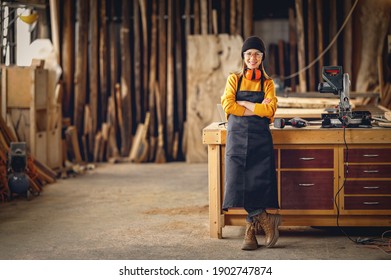 Full body of smiling confident young female joiner in apron standing near workbench and looking at camera friendly while working in craft workshop