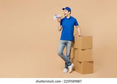 Full body smiling delivery guy employee man wears blue cap t-shirt uniform workwear work as dealer courier stand near stack of cardboard boxes scream in megaphone isolated on plain beige background