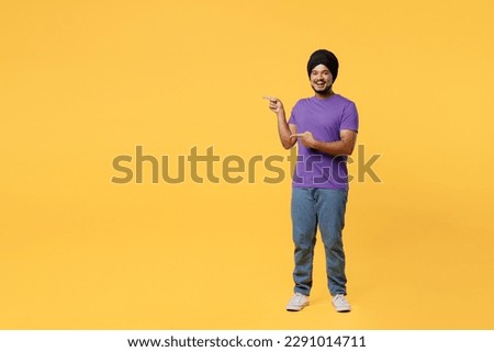 Full body smiling devotee Sikh Indian man ties his traditional turban dastar wear purple t-shirt point index finger aside on workspace area mock up isolated on plain yellow background studio portrait