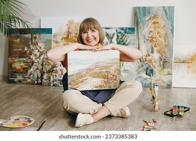 Full body smiling elderly artist woman 50 years old wears casual clothes sit on floor hold in hand show painting on canvas artwork spend free spare time in living room indoor. Leisure hobby concept - Powered by Shutterstock