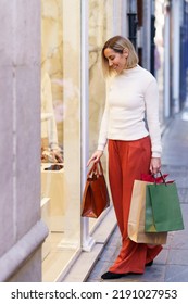 Full body of smiling female customer with purchases in paper shopping bags choosing goods through glass wall of store - Shutterstock ID 2191027953