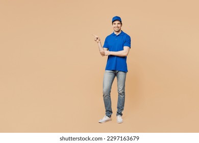 Full body smiling fun delivery guy employee man wear blue cap t-shirt uniform workwear work as dealer courier point index finger aside on area isolated on plain light beige background. Service concept - Powered by Shutterstock