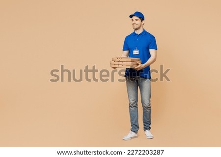 Full body smiling fun delivery guy employee man wear blue cap t-shirt uniform workwear work as dealer courier hold pizza in cardboard flatbox isolated on plain light beige background. Service concept
