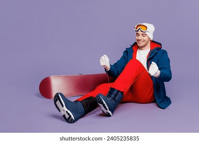 Full body smiling fun man wear blue windbreaker jacket ski goggles mask hat hold snowboard sitting lace up shoes spend extreme weekend winter season in mountains isolated on plain purple background