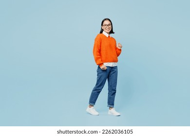 Full body smiling fun young woman of Asian ethnicity wear orange sweater glasses lookiing camera walking strolling isolated on plain pastel light blue cyan background studio. People lifestyle concept