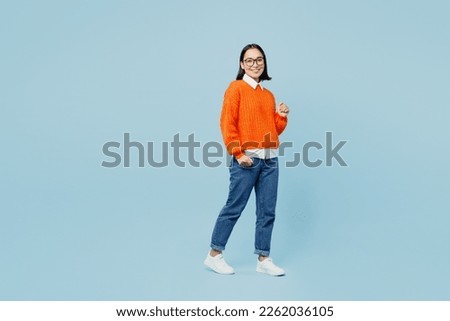 Full body smiling fun young woman of Asian ethnicity wear orange sweater glasses lookiing camera walking strolling isolated on plain pastel light blue cyan background studio. People lifestyle concept