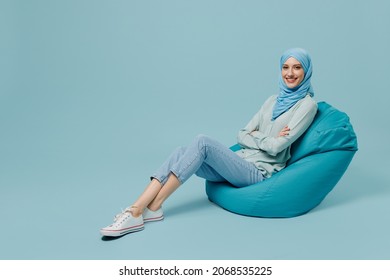 Full Body Smiling Happy Cheerful Young Arabian Asian Muslim Woman In Abaya Hijab Sit In Bag Chair Isolated On Plain Blue Background Studio Portrait. People Uae Middle Eastern Islam Religious Concept