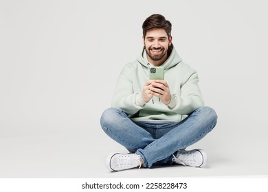 Full body smiling happy cheerful young caucasian man wear mint hoody look camera hold in hand use mobile cell phone isolated on plain solid white background studio portrait. People lifestyle concept - Shutterstock ID 2258228473