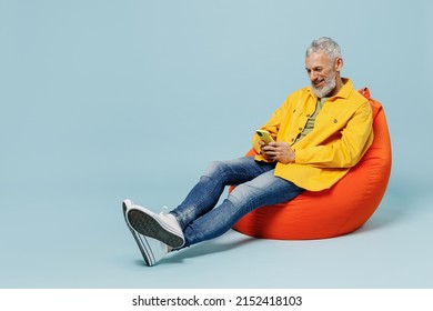 Full body smiling happy elderly gray-haired mustache bearded man 50s in yellow shirt sit in bag chair hold in hand use mobile cell phone isolated on plain pastel light blue background studio portrait. - Powered by Shutterstock