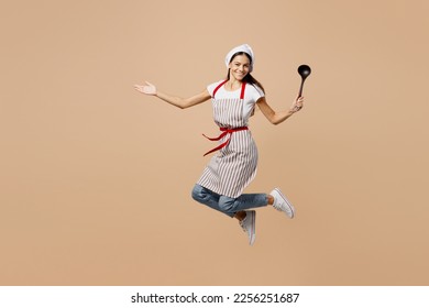 Full body smiling happy fun young housewife housekeeper chef baker latin woman wears apron toque hat jump high hold in hand laddle isolated on plain pastel light beige background. Cook food concept