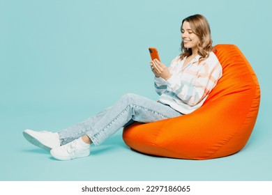 Full body smiling happy fun young woman wear striped hoody sit in bag chair hold in hand use mobile cell phone isolated on plain pastel light blue cyan background studio portrait. Lifestyle concept