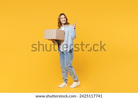 Full body smiling happy fun young woman she wearing blue shirt white t-shirt casual clothes holding cardboard box point finger isolated on plain yellow background studio portrait. Lifestyle concept