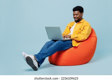 Full body smiling happy young man of African American ethnicity he wear yellow shirt sit in bag chair hold use work on laptop pc computer isolated on plain pastel light blue background studio portrait