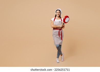 Full body smiling happy young housewife housekeeper chef baker latin woman wear apron toque hat hold in hand frying pan look camera isolated on plain pastel light beige background. Cook food concept - Shutterstock ID 2256251711