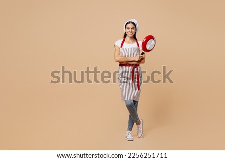 Full body smiling happy young housewife housekeeper chef baker latin woman wear apron toque hat hold in hand frying pan look camera isolated on plain pastel light beige background. Cook food concept
