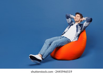 Full body smiling minded happy young man he wear shirt white t-shirt casual clothes sit in bag chair hold hands behind neck isolated on plain blue cyan background studio portrait. Lifestyle concept - Powered by Shutterstock