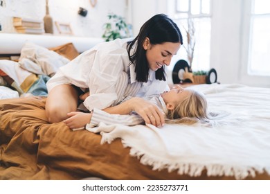 Full body of smiling woman and girl in pajama playing on bed and looking at each other while resting in bedroom at home - Shutterstock ID 2235273771