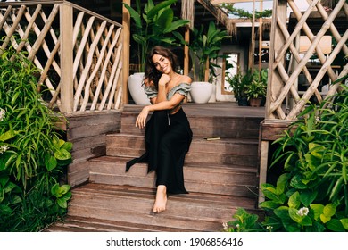 Full Body Smiling Woman In Trendy Outfit Sitting On Steps Of Wooden Terrace Bending Head To Shoulder And Leaning Hands On Knee While Enjoying Plants Decorating Entrance To Summer Cafe Against Plants 