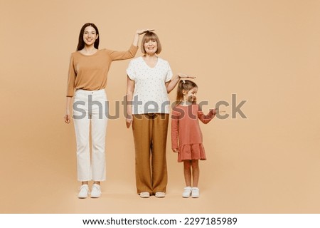 Full body smiling women wearing casual clothes with child kid girl 6-7 years old. Granny mother daughter show height hold hand above head isolated on plain beige background. Family parent day concept