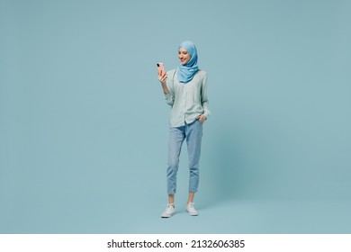 Full body smiling young arabian asian muslim woman in abaya hijab hold in hand use mobile cell phone isolated on plain blue background studio portrait People uae middle eastern islam religious concept
