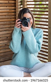 Full Body Of Smiling Young Female Photographer In Casual Outfit Sitting On Wooden Terrace And Taking Pictures With Photo Camera While Enjoying Sunny Weekend